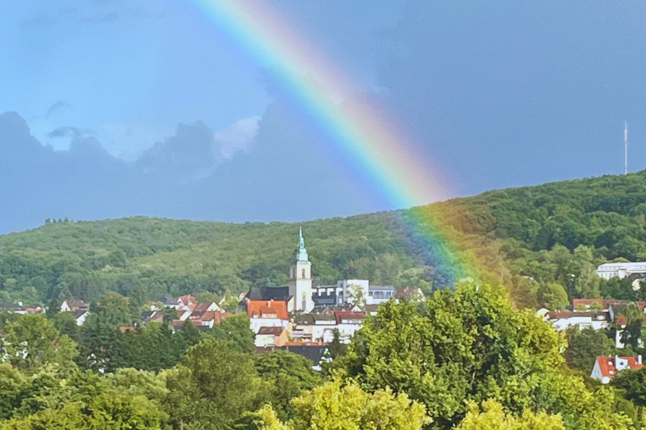 Blick auf die Pfarrkirche Wiebelskirchen. Im Vordergrund grüne Baumkronen, dahinter der Wald, ein graublauer Himmel und darüber ein Regenbogen.