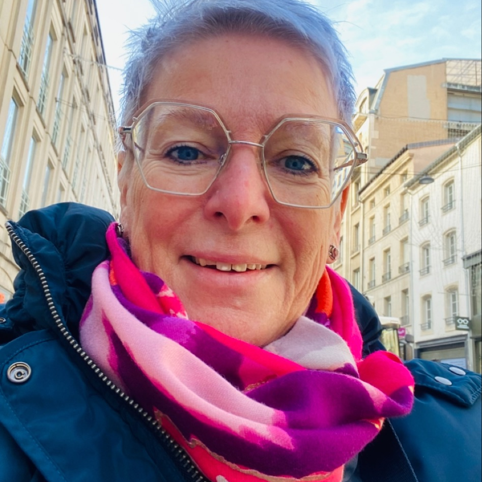 Frau mittleren Alters mit weißen Haaren, buntem Schal in verschiedenen Pink- und Violettönen, mit einer blauen Jacke